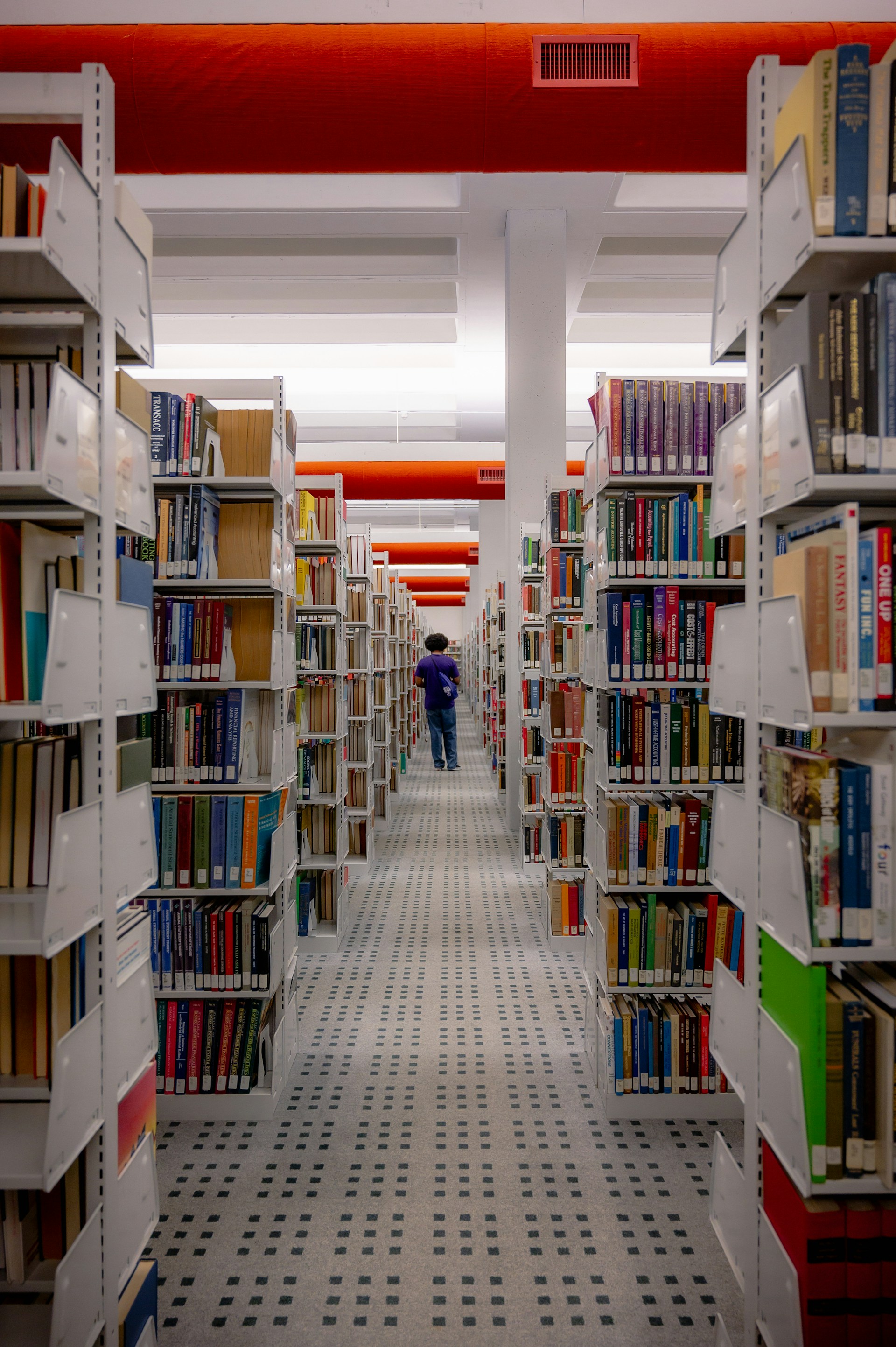 A student walking through a university library aisle lined with bookshelves